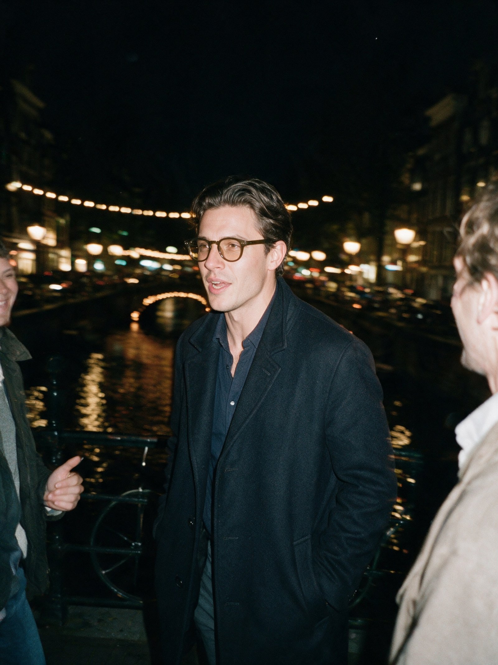 Man crossing a canal bridge at night with friends, flash photo, wearing the olive Bohemian frame